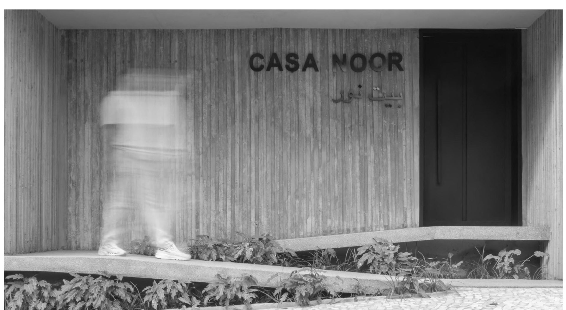 Entrada da Casa Noor, com letreiro em metal sobre parede ripada de madeira